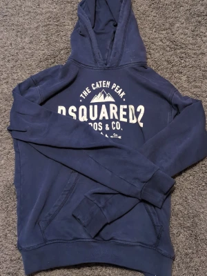 Blå hoodie från Dsquared2 - Mörkblå hoodie från Dsquared2 med vit tryckt logga och text på bröstet. Klassisk känguruficka framtill och huva. Skön bomullsblandning som passar perfekt till chill eller streetwear. Snygg och enkel att matcha med jeans eller joggers. Passar dig mellan 158-168. Pris är ej hugget i sten.