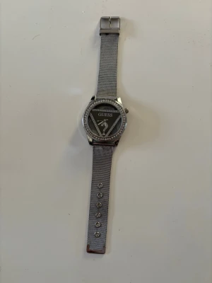 Silverfärgad klocka från Guess - Stilren klocka från Guess med silverfärgat mesharmband och glittrande stenar runt urtavlan. Urtavlan är svart med Guess-logga och detaljer i silver. Perfekt accessoar för att lyfta din outfit.