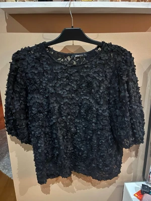 Svart blommig topp från Gina Tricot - Säljer en svart topp från Gina Tricot med 3D-blommor över hela plagget. Toppen har rund halsringning och korta ärmar. Perfekt för dig som vill sticka ut med en unik och trendig look.