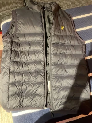 Svart dunväst från Lyle & Scott - Snygg svart dunväst från Lyle & Scott med gul logga på bröstet. Västen har quiltad design med horisontella sömmar och dragkedja framtill. Perfekt för lager-på-lager och enkel att matcha med olika outfits.