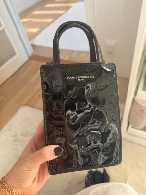 Svart miniväska Karl Lagerfeld - Snygg svart minihandväska från Karl Lagerfeld i glansigt lackmaterial med präglade loggor och motiv. Väskan har två små handtag och en stilren, boxig form. Perfekt för dig som gillar statement-accessoarer och vill ha något trendigt och unikt. Denna design finns inte att hitta längre! Helt ny☺️