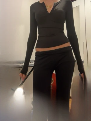 Svart ribbad långärmad topp - Svart långärmad topp med ribbad struktur och v-ringning med knappar framtill. Snygg och slim passform som framhäver siluetten. Perfekt att matcha med jeans eller kjol för en clean look. Jag gillar att ha med yoga pants ELENA GILBERT LOOK använder knappt