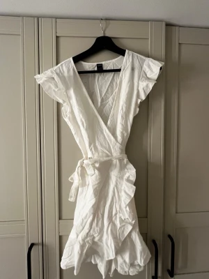 Vit omlottklänning  - Snygg vit kort omlottklänning. Klänningen har volangdetaljer, knytband i midjan och v-ringad hals. Perfekt för varma dagar och somriga tillfällen💕💕