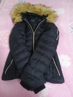 Svart pufferjacka med fuskpäls - Snygg svart pufferjacka från Tally Weijl med guldfärgade dragkedjor och huva med fuskpäls. Jackan har quiltad design, två fickor fram och en extra ficka på ärmen. Perfekt för kalla vinterdagar och ger en trendig look.