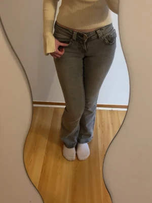 Grå bootcut jeans med låg midja - Säljer ett par grå bootcut jeans med låg midja och klassiska fem fickor. Jeansen har en lätt tvättad look och stängs med dragkedja och knapp. Jag är 157 cm lång och den är lite korta på mig.