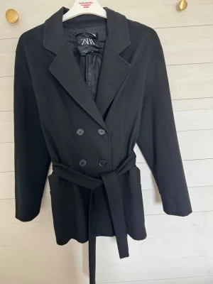 Svart kappa med skärp från Zara - Stilren svart kappa från Zara med dubbelknäppning och tillhörande midjebälte. Klassisk krage, långa ärmar och två fickor framtill. Perfekt för dig som gillar en tidlös look och vill ha en kappa som funkar till det mesta.har ett hål i en av fickorna men inget som märks