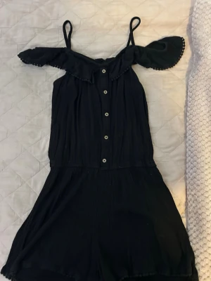 Svart off shoulder jumpsuit - Snygg svart jumpsuit med off shoulder-design och tunna axelband. Knappar framtill och volangdetaljer vid axlarna samt nederkant. Luftigt tyg som känns skönt mot huden, perfekt för varma dagar. Ingen synlig logga eller märke.