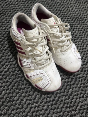 Vita Adidas Adizero sneakers - Snygga vita Adidas Adizero sneakers med lila och beige detaljer. Skorna har klassiska ränder på sidan, ventilerande meshpaneler och robust snörning. Insidan är fodrad i lila tyg och ovandelen är i syntetmaterial och mesh för extra komfort.