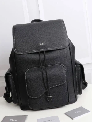 Svart ryggsäck från Dior i läder - Snygg svart ryggsäck från Dior i exklusivt läder med vit kontrastsöm. Ryggsäcken har ett stort lock med dragsko, en framficka och två sidofickor för extra förvaring. Minimalistisk design med diskret Dior-logga på locket. Perfekt för dig som vill ha både stil och funktion.