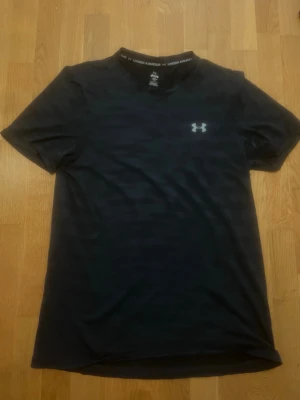Svart träningströja från Under Armour - Snygg svart kortärmad träningströja från Under Armour med diskret mönster och logga på bröstet. Tillverkad i ett lätt och snabbtorkande funktionsmaterial som passar perfekt för träning och sport.