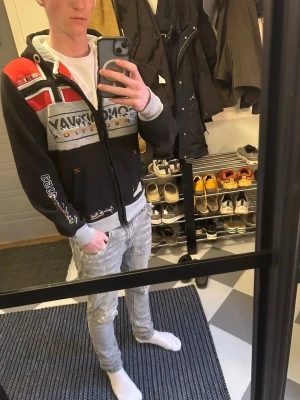 Svart hoodie från Geographical Norway - Säljer en svart hoodie från Geographical Norway i storlek s med dragkedja, broderade detaljer och tryck i vitt, grått och rött. Hoodien har huva med snörning och mönster med norska flaggan på bröstet. Perfekt för dig som gillar streetwear och snygga detaljer.
