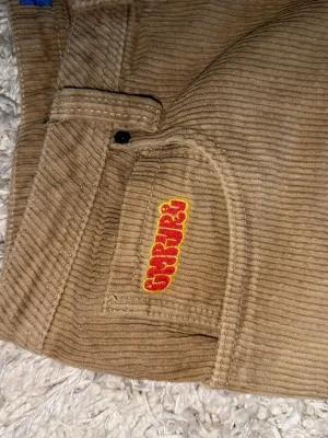 Beiga corduroybyxor Gramicci - Säljer ett par beiga manchesterbyxor från empyre med klassisk femficksdesign och broderad logga i rött och gult på fickan. Byxorna har raka ben och är tillverkade i mjuk, bredspårig manchester som ger en avslappnad och trendig look.