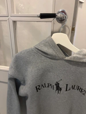 Grå hoodie från Ralph Lauren - Snygg grå hoodie från Ralph Lauren med klassisk logga och tryck framtill. Den är tyvärr klippt längst ner och lite knott, klippningen går att fixa!! Hoodien har huva och är tillverkad i mjukt, fleeceliknande material som känns riktigt skönt mot huden. Perfekt för en avslappnad och stilren look.