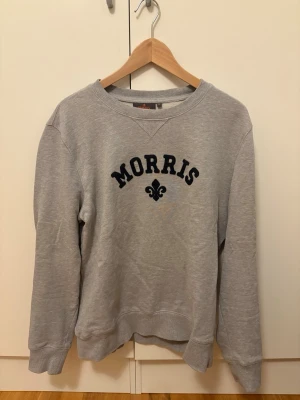 Grå sweatshirt från Morris - Grå sweatshirt från Morris med svart broderad logga och emblem framtill. Klassisk rund halsringning och långa ärmar. Mjuk och bekväm modell i bomull, perfekt för en avslappnad stil.