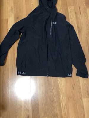 Svart hoodie från Under Armour - Svart hoodie från Under Armour med dragkedja framtill och huva med dragsko. Diskret logga på bröstet och tryck längs nederkanten. Tillverkad i ett mjukt och sportigt material, perfekt för en aktiv livsstil.