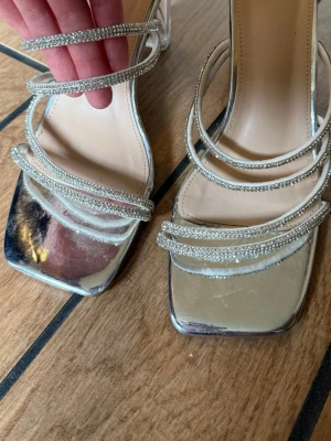 Silvriga klackar med strass  - Glittriga silvriga klackskor med fyrkantig tå, flera tunna remmar dekorerade med strass och transparent blockklack. Skorna har metallic-finish och insida i beige syntetmaterial. Perfekt för dig som vill sticka ut med din stil. Använda 1 gång.