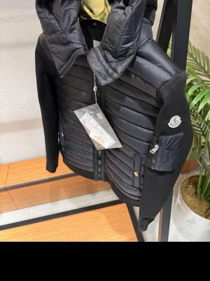 Svart pufferjacka från Moncler - Snygg svart pufferjacka från Moncler med quiltad front och huva. Ärmarna är i stickat material och har en ficka med dragkedja samt Moncler-logga på vänster arm. Jackan har två framfickor med tryckknappar och dragkedja framtill.