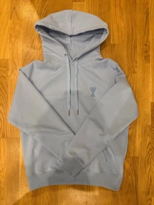 Ljusblå hoodie från Ami Paris - Snygg ljusblå hoodie från Ami Paris med det ikoniska hjärta-A broderiet på bröstet. Hoodien har dragsko i huvan, ribbade muddar och en klassisk känguruficka. Tillverkad i mjuk bomull för maximal komfort.