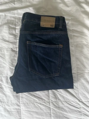 Mörkblå jeans från 157 - Säljer ett par klassiska mörkblå jeans från 157 med raka ben och kontrastsömmar. Jeansen har en bakficka och en snygg läderpatch i midjan. Perfekta för en stilren och enkel look.