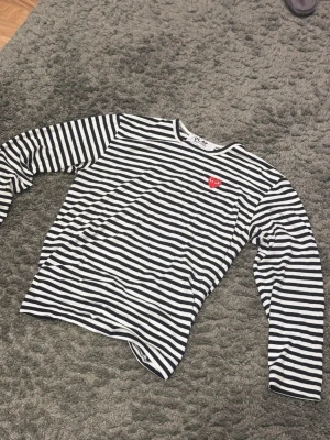Randig långärmad topp Comme des Garçons - Säljer en svartvit randig långärmad topp från Comme des Garçons Play med det ikoniska röda hjärtat broderat på bröstet. Toppen har rund halsringning och är tillverkad i mjuk bomull. Perfekt för en stilren och trendig look.