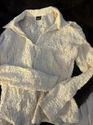  crinkle-blus från Gina Tricot - Trendig kräm vit blus från Gina Tricot med crinkle-effekt och klassisk krage. Blusen har lång ärm och knäppning framtill. Materialet är lätt och stretchigt för extra komfort.