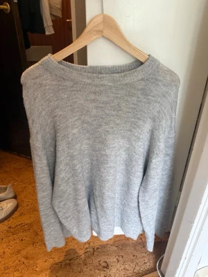 Ljusgrå stickad tröja 157 - Mysig ljusgrå stickad tröja från 157 med rund halsringning och avslappnad passform. Perfekt för lager-på-lager och enkel att matcha med jeans eller kjol. Tröjan har långa ärmar och ribbstickade detaljer vid halsen.