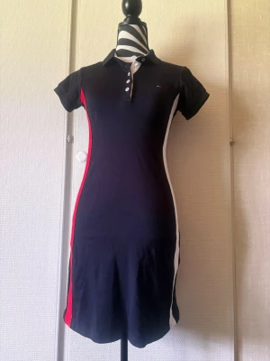 Marinblå kort klänning Tommy Hilfiger - Sportig marinblå kort klänning från Tommy Hilfiger med polokrage, knappar framtill och kontrasterande vita och röda paneler längs sidorna. Klänningen har korta ärmar och är tillverkad i mjuk bomull för en bekväm passform.