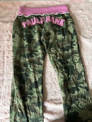 Camo mjukisbyxor från Paul Frank - Gröna camo-mönstrade mjukisbyxor från Paul Frank med rosa resår i midjan och stora rosa bokstäver bak. Byxorna har det klassiska Julius-apan-trycket över hela tyget och en avslappnad passform. Perfekta för en chill och lekfull stil.