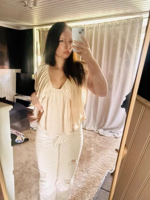 Beige omlottopp med volang - Snygg beige omlottopp med djup v-ringning och volangdetaljer. Toppen är ärmlös och har en luftig passform som ger en avslappnad och trendig look. Perfekt att styla med jeans eller kjol för en cool vardagsstil.