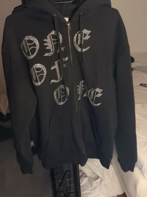 Svart rhinestone hoodie - Svart zip-hoodie med huva och dragsnören. Framsidan har ett stort tryck i glittrande strass-stenar med texten 'ONE OF ONE' i gotisk stil. Mjuk bomullskänsla och klassisk passform, perfekt för en streetwear-look.