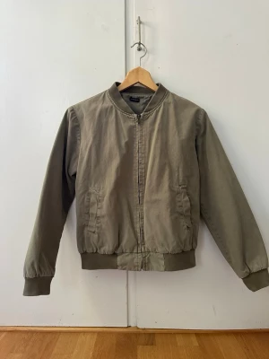 Grön bomberjacka  - Snygg olivgrön bomberjacka från Brandy Melville med klassisk ribbad krage, muddar och midja. Jackan har dragkedja framtill och två fickor med lock. Tillverkad i ett mjukt bomullsmaterial som ger en avslappnad look.