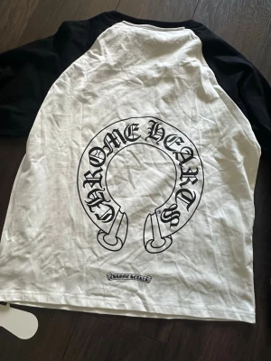 Chrome Hearts x Comme des Garçons t-shirt - Snygg t-shirt i samarbete mellan Chrome Hearts och Comme des Garçons. Vit bas med svarta raglanärmar och stor Chrome Hearts-logga på ryggen. Tillverkad i mjuk bomull, perfekt för en streetwear-look.