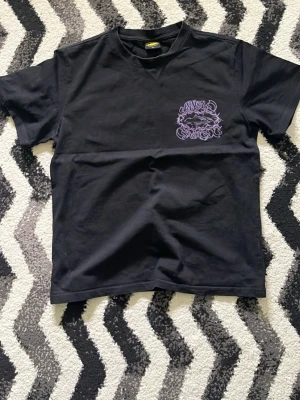 Svart t-shirt  - Cool svart t-shirt med ett unikt lila tryck på bröstet. T-shirten har klassisk passform och rund halsringning. Perfekt för dig som gillar streetwear och vill sticka ut med en snygg detalj. Passar även S