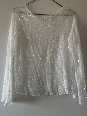 Vit stickad tröja med paljetter - Säljer en vit stickad tröja med långa ärmar och rund hals. Tröjan är dekorerad med glittrande paljetter över hela ytan vilket ger en festlig och trendig look. Materialet är tunt och lätt, perfekt för att sticka ut i mängden.