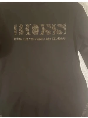 Svart BOSS t-shirt med paljettdetalj - Snygg svart t-shirt från BOSS med coolt paljettmönster på bröstet. T-shirten har klassisk passform och rund halsringning. Perfekt för dig som vill sticka ut med en stilren men ändå unik look.
