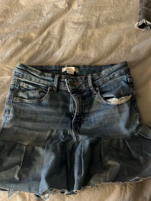 Blå jeanskjol med volangkant - Snygg blå jeanskjol med volangkant nertill och råa kanter. Kjolen har klassiska jeansfickor fram och bak, bälteshällor och knappstängning. Perfekt för dig som gillar en avslappnad och trendig stil. Kjolen är egensydd!! 