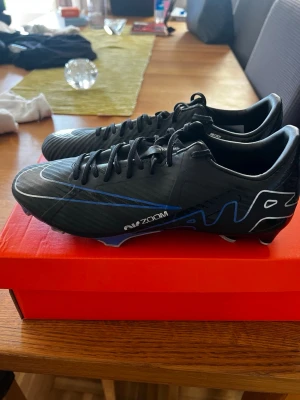 Nike Mercurial Zoom svart/blå - Säljer ett par Nike Mercurial Zoom fotbollsskor i svart med blå och vita detaljer. Skorna har en modern, strömlinjeformad design med snörning och syntetiskt material för bra grepp och känsla på planen. Perfekta för dig som vill ha stilrena och snabba skor.