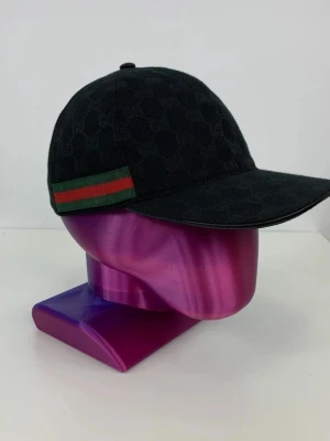 Svart keps från Gucci med logomönster - Svart keps från Gucci med diskret GG-logomönster över hela ytan. Klassisk böjd skärm och ikoniskt grönt och rött band på sidan. Tillverkad i canvasmaterial som ger en lyxig känsla och snygg passform.