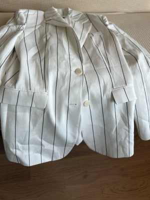 Vit kavaj med svarta ränder från Mango - Snygg vit kavaj från Mango med svarta vertikala ränder. Klassisk passform med två knappar framtill, fickor med lock och krage. Perfekt för en trendig och stilren look. Materialet känns lätt och passar bra till många outfits.