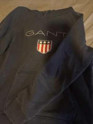 Mörkblå hoodie från GANT - Snygg mörkblå hoodie från GANT med broderad logga och amerikanskinspirerat sköldmärke på bröstet. Hoodien har en klassisk känguruficka framtill och ribbade muddar vid ärmslut och nederkant. Perfekt för en avslappnad och stilren look.