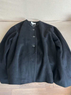 Svart klassisk ullrock med sjal  - Stilren svart rock med rak passform och knäppning framtill med fyra knappar. Tillverkad i ett tjockare ullmaterial som passar perfekt för kyligare dagar. Enkel design utan synliga fickor eller detaljer, vilket ger en tidlös look.