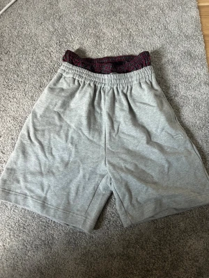 Grå mjukisshorts med resårmidja - Grå mjukisshorts med elastisk midja och diskret broderad logga på benet. Insidan av midjan har ett röd- och svartmönstrat tyg. Perfekta för chill eller träning, tillverkade i mjuk bomull.