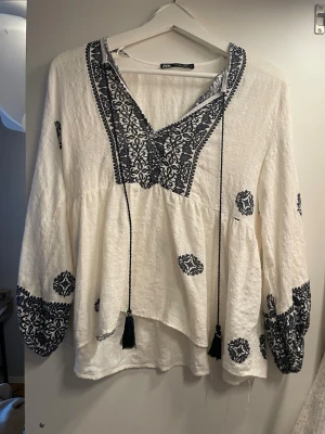 Vit boho-blus med brodyr från Zara - Vit bohemisk blus från Zara med blå broderi längs v-ringningen, ärmsluten och utspritt över hela plagget. Blusen har långa, vida ärmar och dekorativa tofsar vid halsen. Tillverkad i ett lätt, strukturerat tyg som ger en avslappnad känsla.