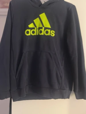 Svart hoodie från Adidas med logga - Svart hoodie från Adidas med stor neongul logga och text på bröstet. Klassisk känguruficka framtill och huva. Mjuk bomullsblandning som är skön att ha på sig.