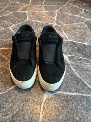 Svarta axel Arigato laceless  - Stilrena svarta sneakers från Axel Arigato med slip-on design och vit sula. Ovandelen är i mocka och textil, med diskret logga på plösen. Perfekta för en clean och modern look.