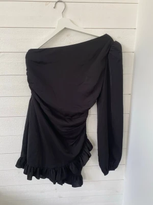 Svart oneshoulder klänning med volang - Trendig svart kortklänning med oneshoulder-design och lång ärm. Klänningen har snygga rynkade detaljer och volang längst ner för en cool look. Perfekt för dig som gillar att sticka ut med en unik stil.
