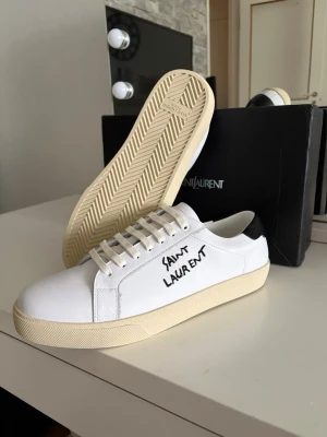 Saint Laurent vita sneakers med svart detalj - Snygga vita sneakers från Saint Laurent med svart hälparti och handskriven logga på sidan. Skorna har en klassisk låg profil, vita snören och en beige sula. Tillverkade i läder för en lyxig känsla och stilren look.