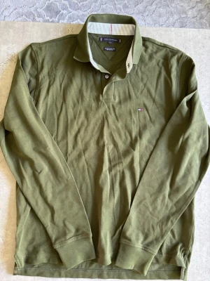 Olivgrön långärmad pikétröja Tommy Hilfiger - Snygg olivgrön långärmad pikétröja från Tommy Hilfiger med klassisk krage och knappslå. Liten broderad logga på bröstet och ribbade muddar vid ärmslut. Perfekt för en avslappnad men stilren look.