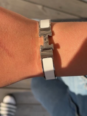 Vitt H-armband i metall - Snyggt armband från HM silverfärgad metall med vita emaljdetaljer. Armbandet har den ikoniska H-formade låset och en stilren, modern design som passar perfekt till alla outfits. Enkel att matcha och ger en lyxig touch till din stil.