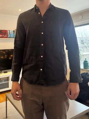 Svart skjorta i bomull med knappar - Snygg svart skjorta i bomull med klassisk krage och vita knappar hela vägen framtill. Skjortan har lång ärm och en avslappnad passform, perfekt för en stilren look.